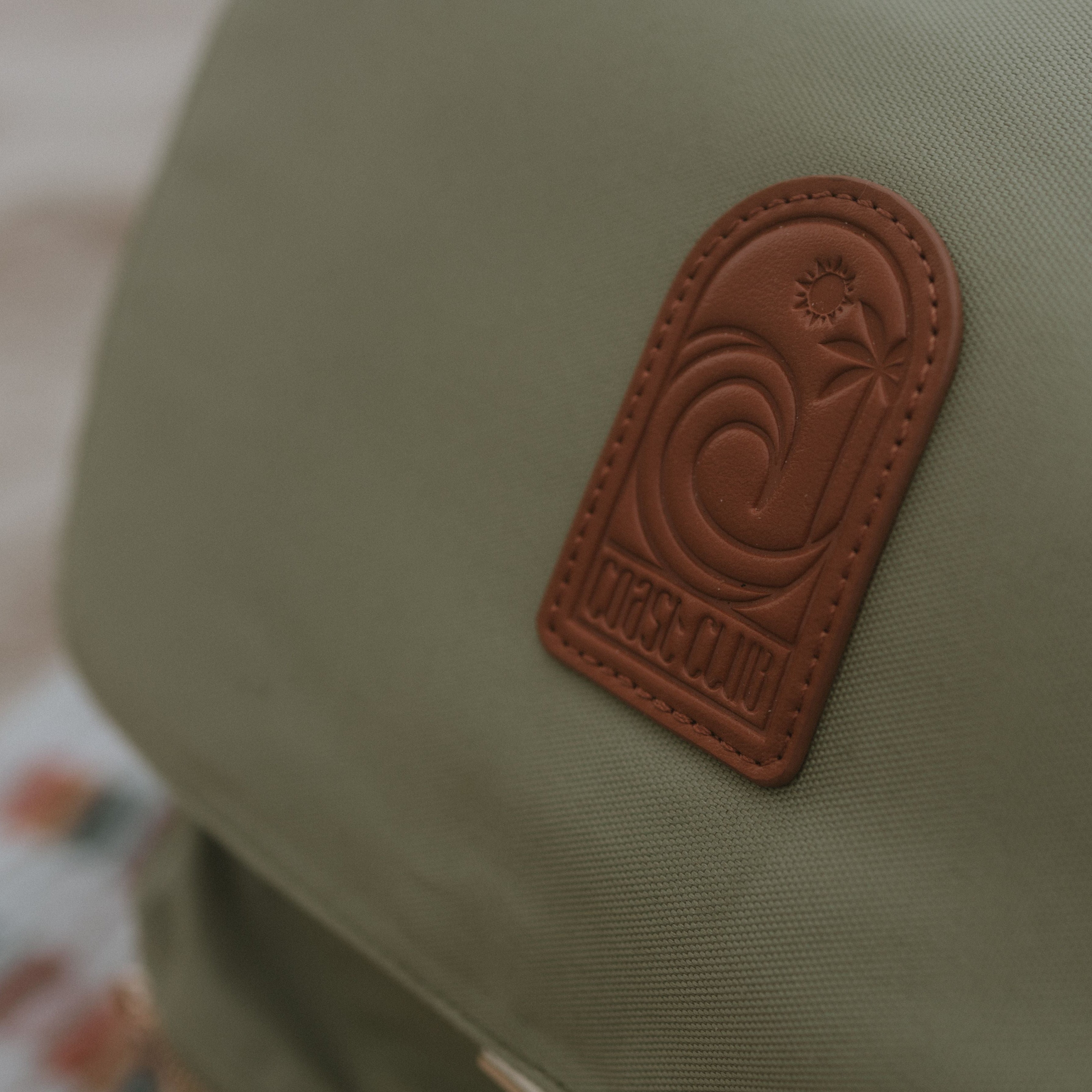 Close-up of a sage bag with a brown leather patch featuring a logo and text.