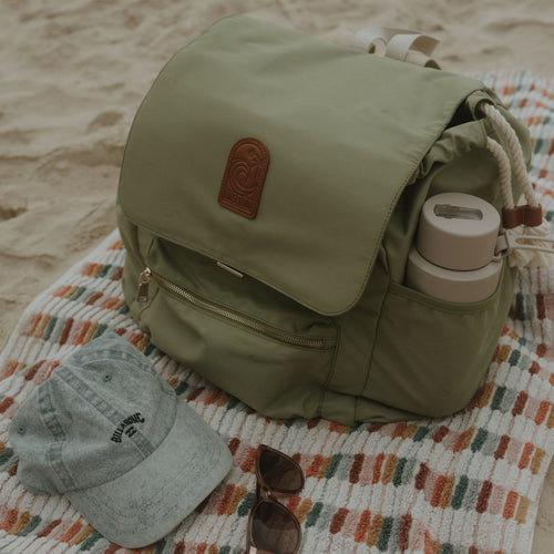 Sage oversized backpack with a bottle, sunglasses, and cap on a patterned blanket on sand