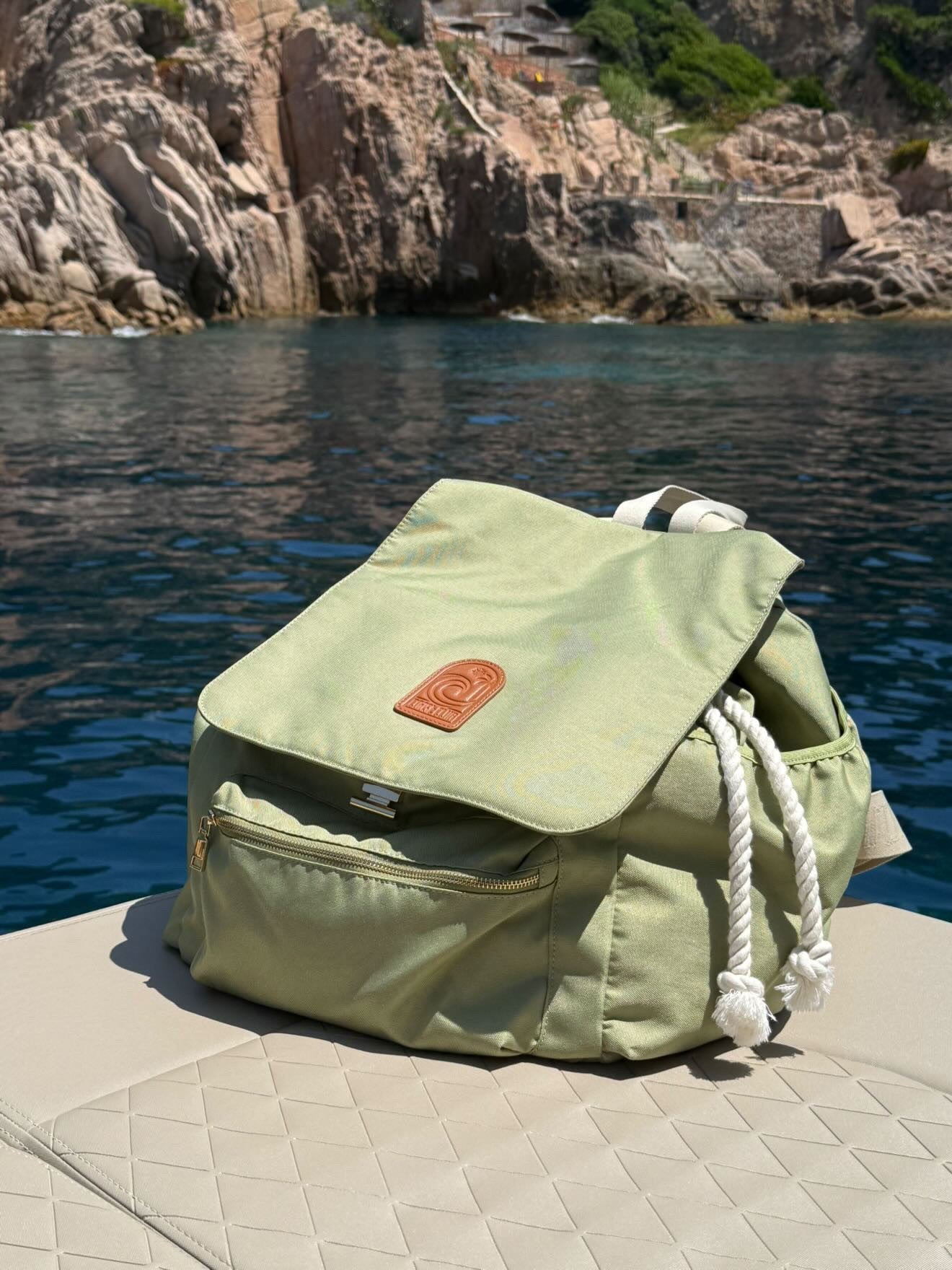 A sage green large backpack with a brown leather patch and white rope drawstrings sits on a beige boat deck with a rocky sea cliff in the background.