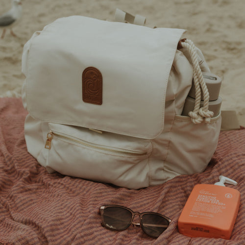 Beige oversized backpack on a beach with sunglasses and sunscreen