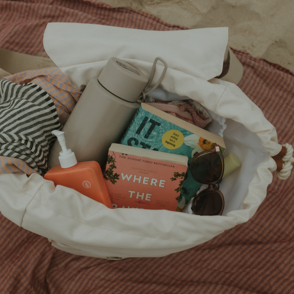 Open bag with travel items including a thermos, books, and sunglasses on a beach towel